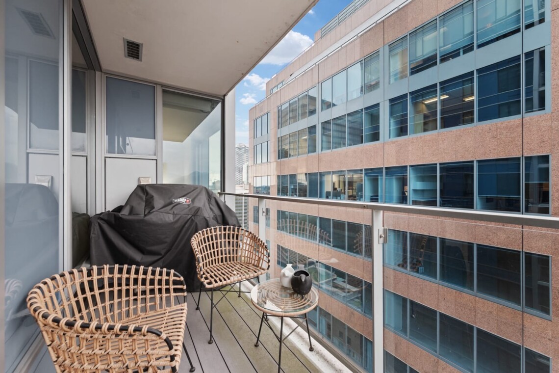 Balcony with wicker chairs and a small glass table overlooking a tall office building with glass windows.