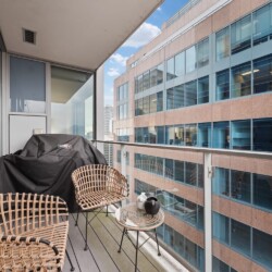 Balcony with wicker chairs and a small glass table overlooking a tall office building with glass windows.