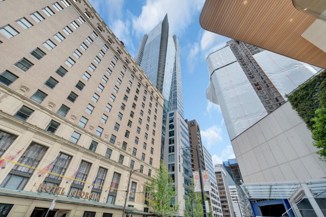 Tall city street with a beige historic hotel Georgia on the left and modern glass towers towering overhead against a blue sky.