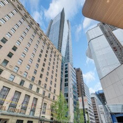 Tall city street with a beige historic hotel Georgia on the left and modern glass towers towering overhead against a blue sky.