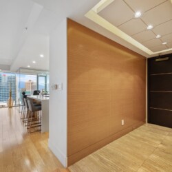 Modern apartment entry with a dark wood door, warm wood accent wall, and city skyline visible through large windows.