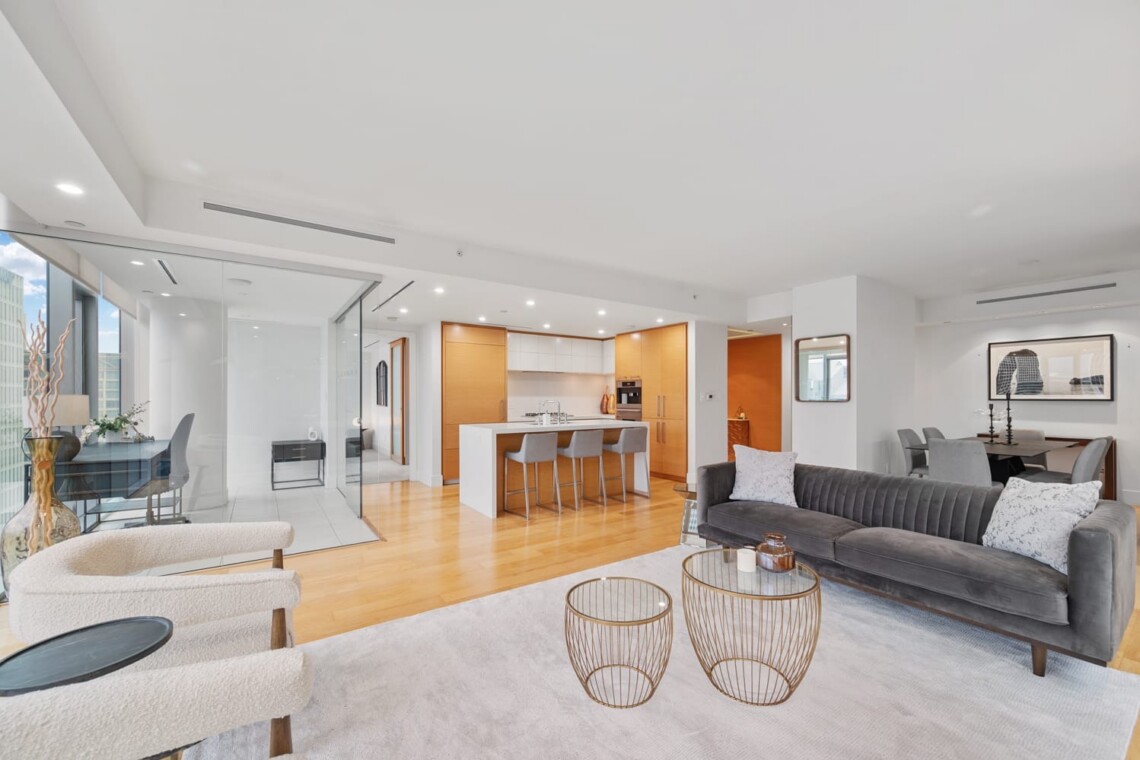 Open-concept living space with a gray sofa, two round wire coffee tables, and cream armchairs facing a bright kitchen island, dining area in back, and glass partition.”,