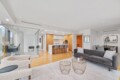 Open-concept living space with a gray sofa, two round wire coffee tables, and cream armchairs facing a bright kitchen island, dining area in back, and glass partition.”,