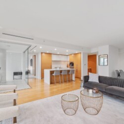 Open-concept living space with a gray sofa, two round wire coffee tables, and cream armchairs facing a bright kitchen island, dining area in back, and glass partition.”,