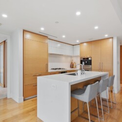 Open-plan kitchen with a white island, wooden cabinets, and stainless appliances; bar seating along island with gray stools.