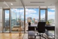 Corner office with a dark desk and chair, overlooking a city skyline through floor-to-ceiling windows.
