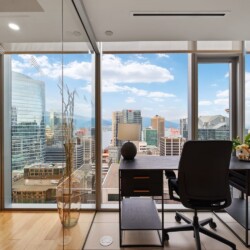 Corner office with a dark desk and chair, overlooking a city skyline through floor-to-ceiling windows.