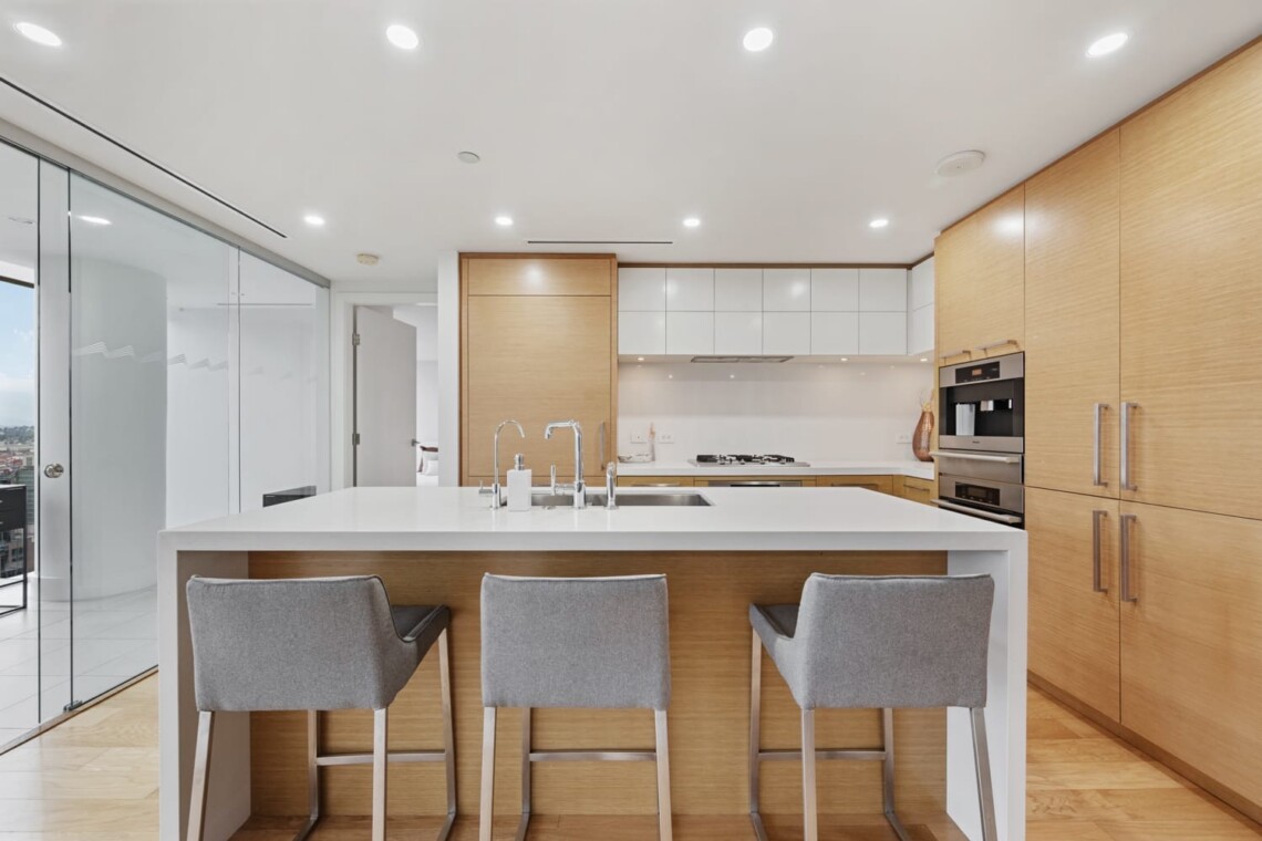Modern kitchen with a white island, gray upholstered stools, and warm wood cabinetry; built-in ovens on the right and a glass door to a balcony on the left.