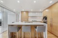 Modern kitchen with a white island, gray upholstered stools, and warm wood cabinetry; built-in ovens on the right and a glass door to a balcony on the left.