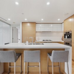 Modern kitchen with a white island, gray upholstered stools, and warm wood cabinetry; built-in ovens on the right and a glass door to a balcony on the left.