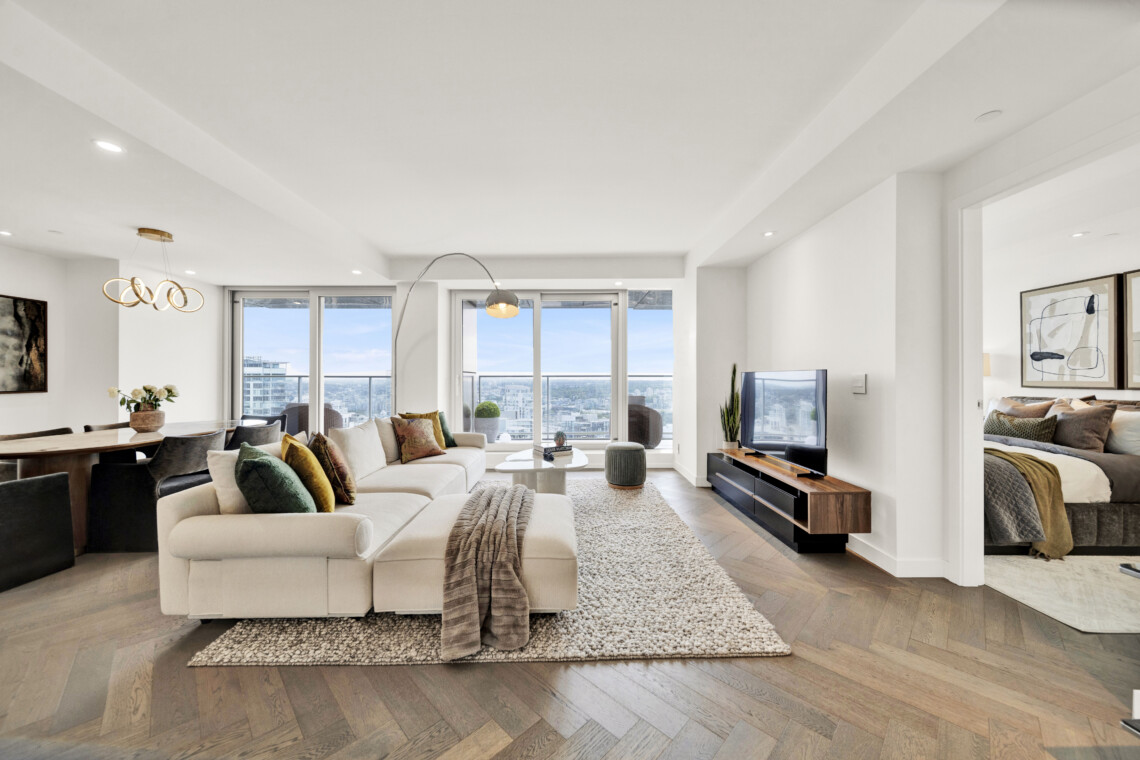 Bright, open living room with a large beige sectional, colorful pillows, and a white rug facing a city view through floor-to-ceiling windows.