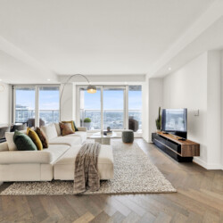 Bright, open living room with a large beige sectional, colorful pillows, and a white rug facing a city view through floor-to-ceiling windows.