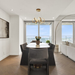 Modern dining area with a long white table, dark chairs, and a gold loop chandelier, overlooking a cityscape through large windows.