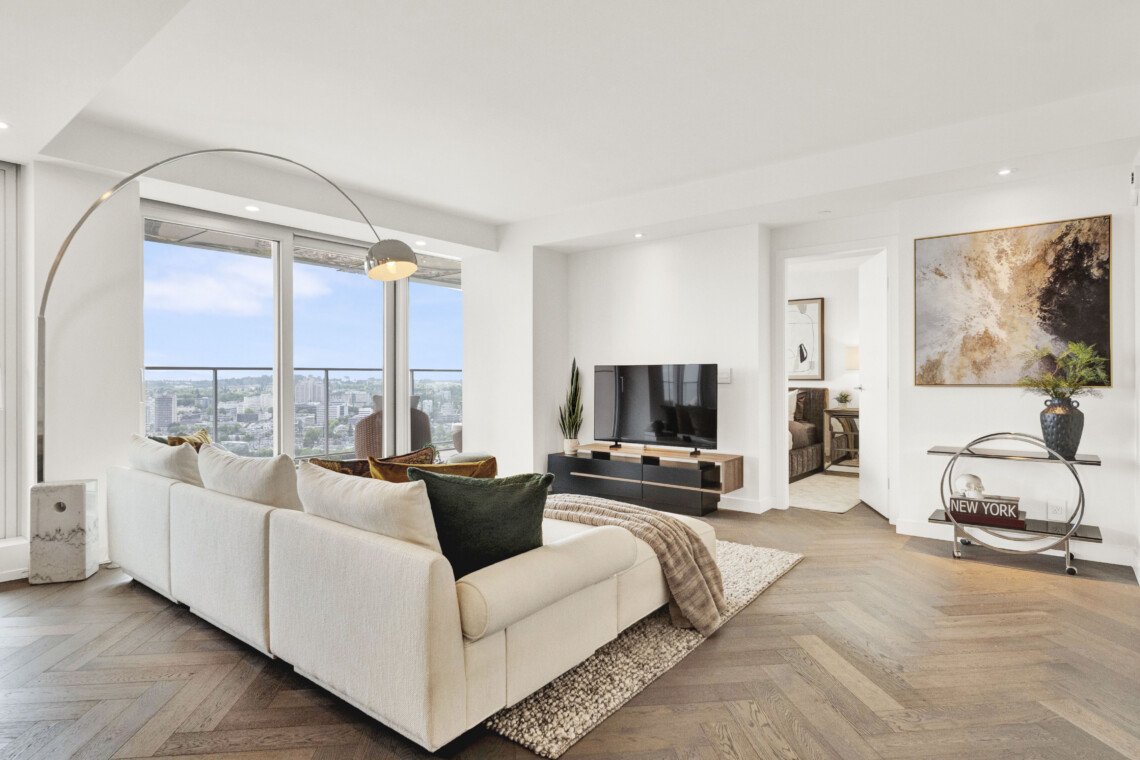 Bright contemporary living room with a white sectional sofa, arc floor lamp, and large windows showing a city skyline. Abstract wall art hangs on the right.
