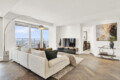 Bright contemporary living room with a white sectional sofa, arc floor lamp, and large windows showing a city skyline. Abstract wall art hangs on the right.