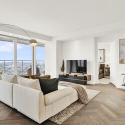 Bright contemporary living room with a white sectional sofa, arc floor lamp, and large windows showing a city skyline. Abstract wall art hangs on the right.