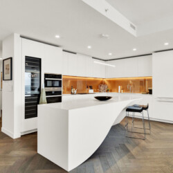 White modern kitchen with a curved island, orange under-cabinet lighting, and built-in ovens in a tall cabinet.