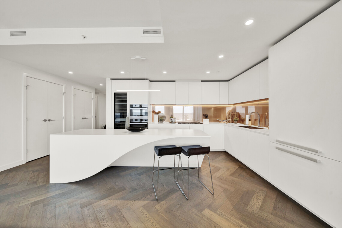 Contemporary white kitchen with a curved island, sleek white cabinets, and warm under-cabinet lighting over a wooden herringbone floor.