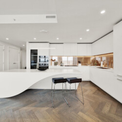 Contemporary white kitchen with a curved island, sleek white cabinets, and warm under-cabinet lighting over a wooden herringbone floor.