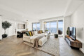 Bright, modern living room with a beige sectional, colorful throw pillows, and large windows overlooking the city skyline.
