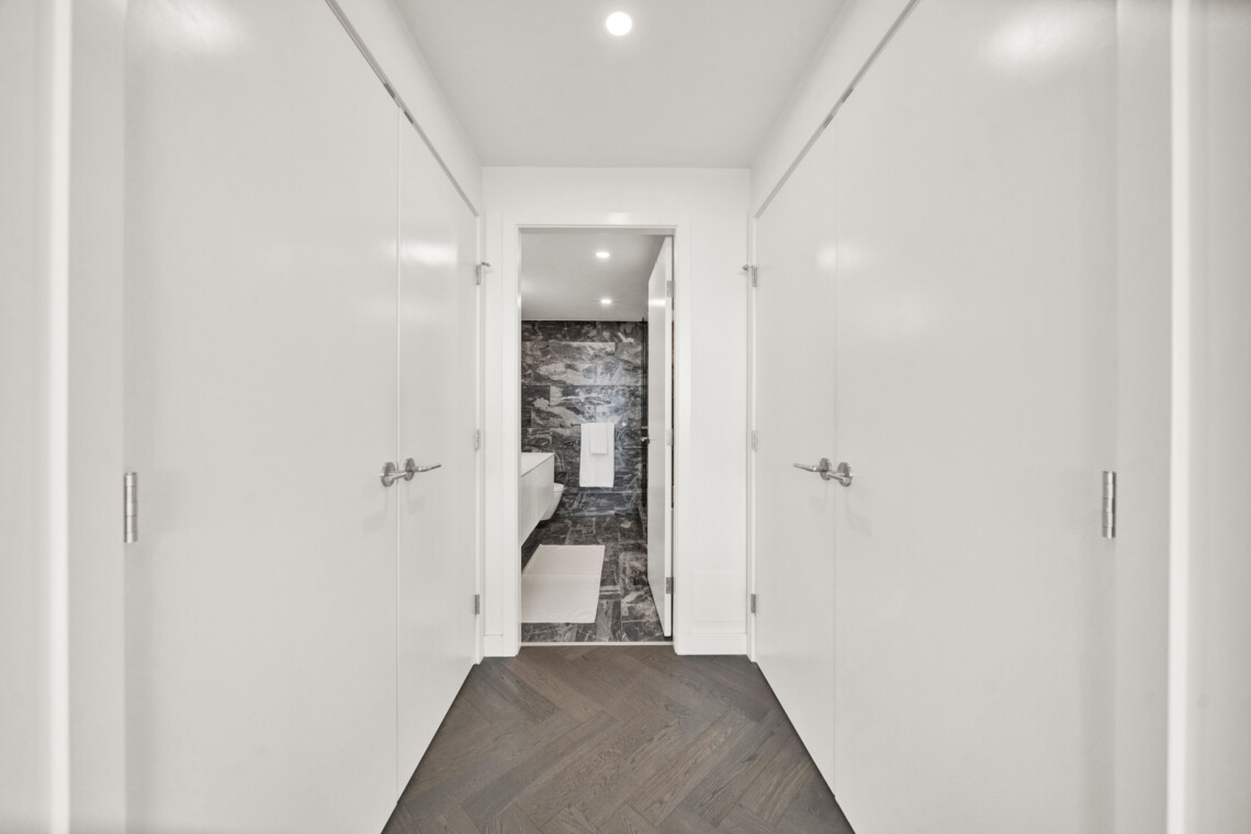 Narrow white hallway lined with doors on both sides, opening into a bathroom with dark marble tiles at the far end.