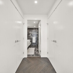 Narrow white hallway lined with doors on both sides, opening into a bathroom with dark marble tiles at the far end.