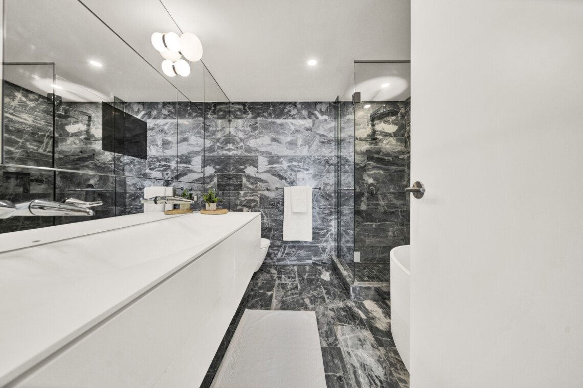 Modern bathroom with a white vanity, marble walls and floor, and a glass-enclosed shower; white towels and small green plants on the counter add accents.