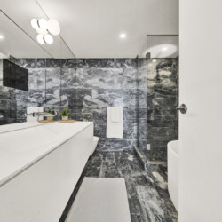 Modern bathroom with a white vanity, marble walls and floor, and a glass-enclosed shower; white towels and small green plants on the counter add accents.