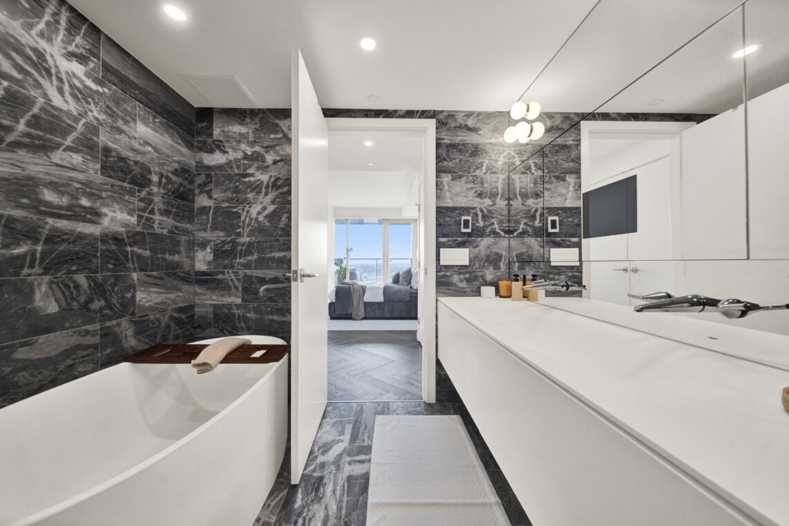 Luxurious white bathroom with dark marble walls, white cabinetry, and a bathtub along the left edge; doorway opens to a bright living area.