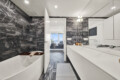 Luxurious white bathroom with dark marble walls, white cabinetry, and a bathtub along the left edge; doorway opens to a bright living area.