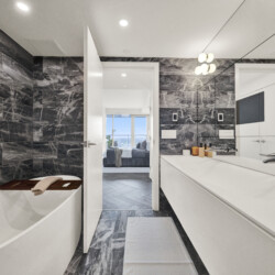 Luxurious white bathroom with dark marble walls, white cabinetry, and a bathtub along the left edge; doorway opens to a bright living area.