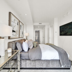 Modern hotel-style bedroom with a gray upholstered bed, abstract art on the wall, and a wall-mounted TV on the right.