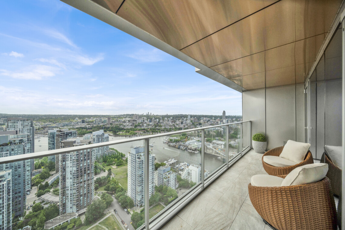 Balcony seating with wicker chairs overlooking a city river and skyline under a blue sky, modern glass railing in the foreground.