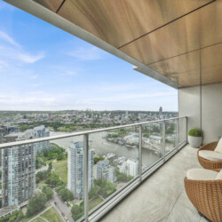 Balcony seating with wicker chairs overlooking a city river and skyline under a blue sky, modern glass railing in the foreground.