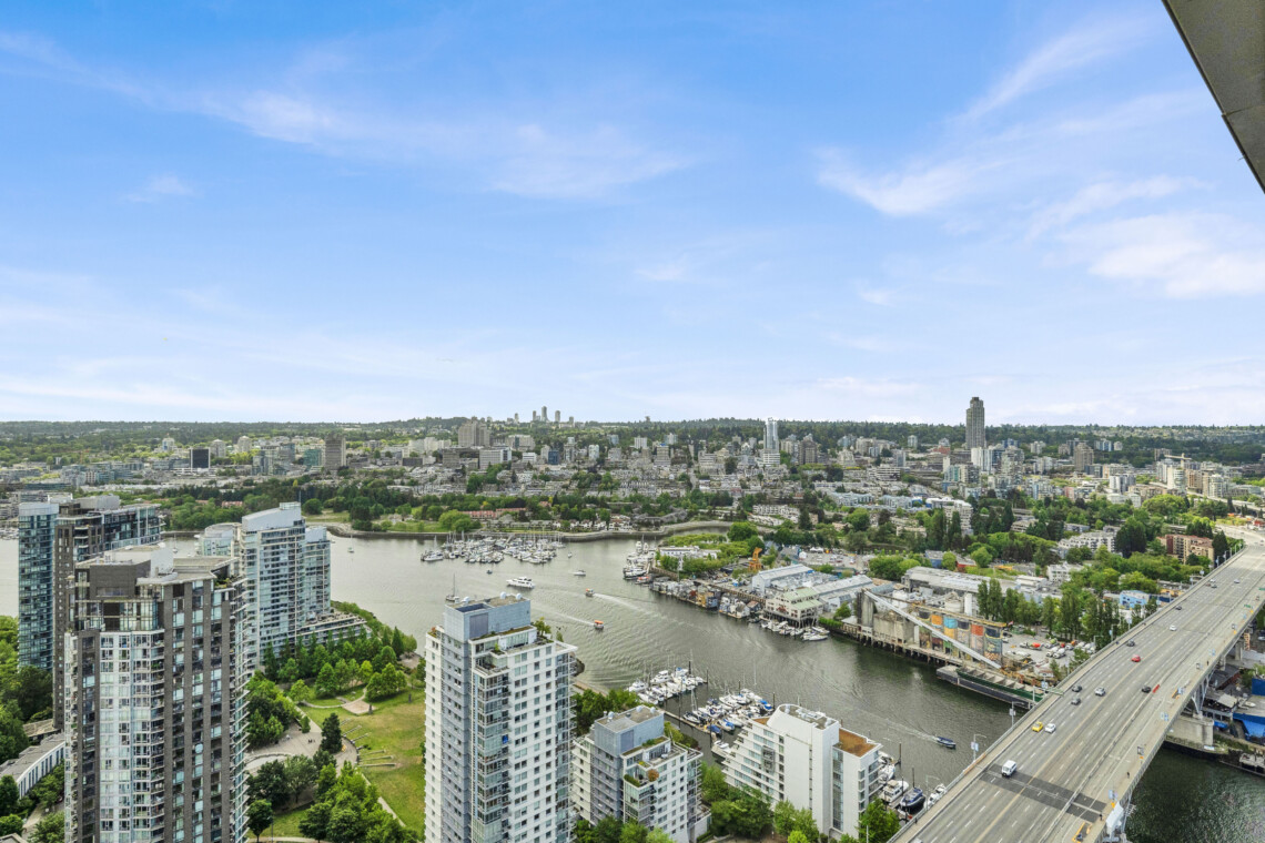 Aerial view of a modern city along a river, with tall residential towers and a nearby marina under a blue sky, right-side highway visible.