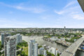 Aerial view of a modern city along a river, with tall residential towers and a nearby marina under a blue sky, right-side highway visible.