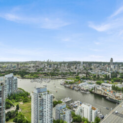 Aerial view of a modern city along a river, with tall residential towers and a nearby marina under a blue sky, right-side highway visible.