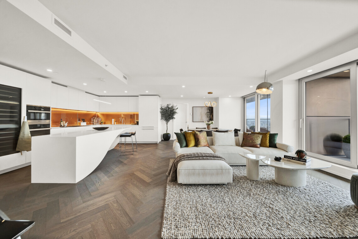 Open-concept living area with a white kitchen island, orange-lit backsplash, and a cream sofa with colorful cushions facing large windows.
