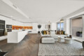 Open-concept living area with a white kitchen island, orange-lit backsplash, and a cream sofa with colorful cushions facing large windows.