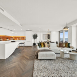 Open-concept living area with a white kitchen island, orange-lit backsplash, and a cream sofa with colorful cushions facing large windows.