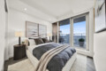 Modern bedroom with a plush upholstered bed, gray and beige bedding, two nightstands with lamps, and large glass doors opening to a balcony with a city view.