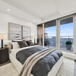 Modern bedroom with a plush upholstered bed, gray and beige bedding, two nightstands with lamps, and large glass doors opening to a balcony with a city view.