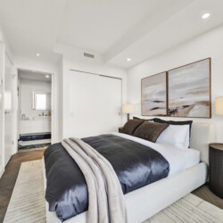 Bright bedroom with a low bed, dark gray bedding, and a cozy beige throw; bedside lamp and round black nightstand on the right; two landscape paintings above the bed.