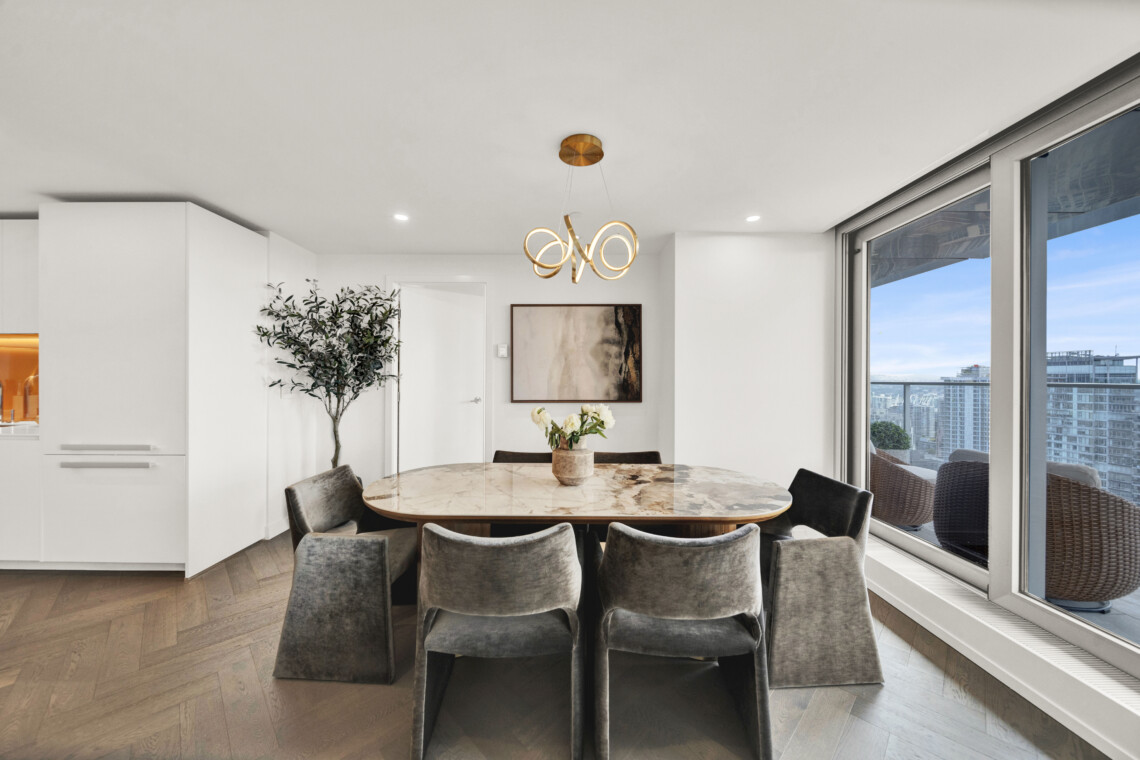 Round marble dining table with gray upholstered chairs in a bright modern dining area; a gold looping chandelier hangs above, with a city skyline visible through large windows.