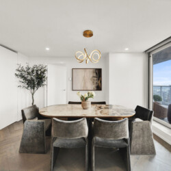 Round marble dining table with gray upholstered chairs in a bright modern dining area; a gold looping chandelier hangs above, with a city skyline visible through large windows.