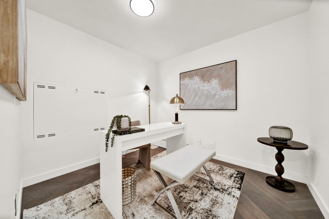 Bright white home office corner with a white desk, bench seating, a potted plant, and a large framed wave artwork on the wall; warm lighting from a brass floor lamp.
