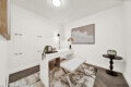 Bright white home office corner with a white desk, bench seating, a potted plant, and a large framed wave artwork on the wall; warm lighting from a brass floor lamp.