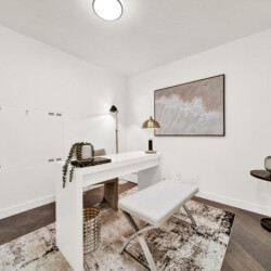 Bright white home office corner with a white desk, bench seating, a potted plant, and a large framed wave artwork on the wall; warm lighting from a brass floor lamp.