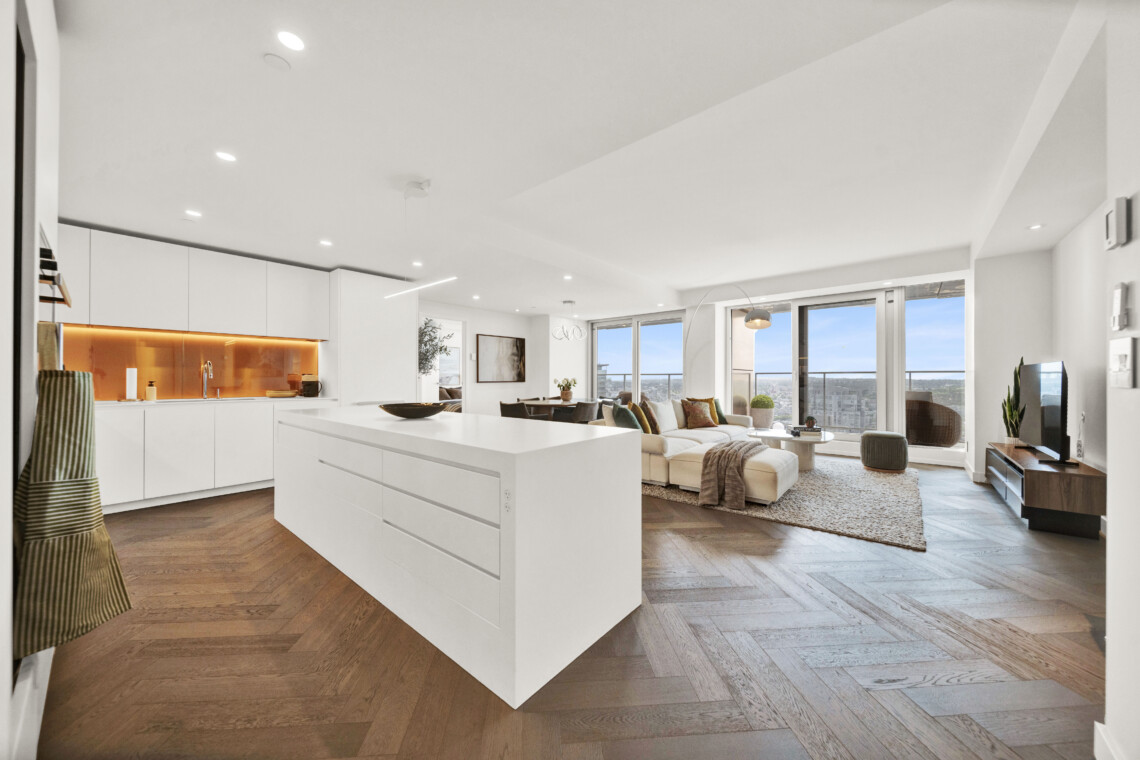 Open-plan kitchen and living area with white cabinets, a large white island, orange backsplash, and a seating area by windows with a city view.
