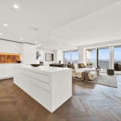 Open-plan kitchen and living area with white cabinets, a large white island, orange backsplash, and a seating area by windows with a city view.